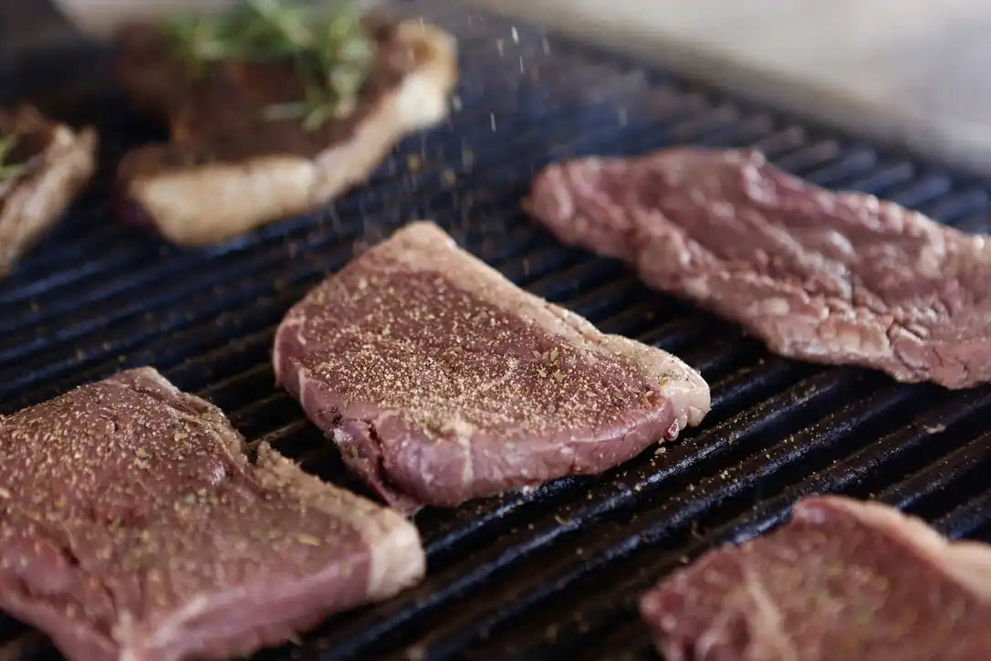 Raw, seasoned beef steaks on a hot grill beginning to sear, highlighting the freshness and preparation of Drummond Ranch premium cuts.