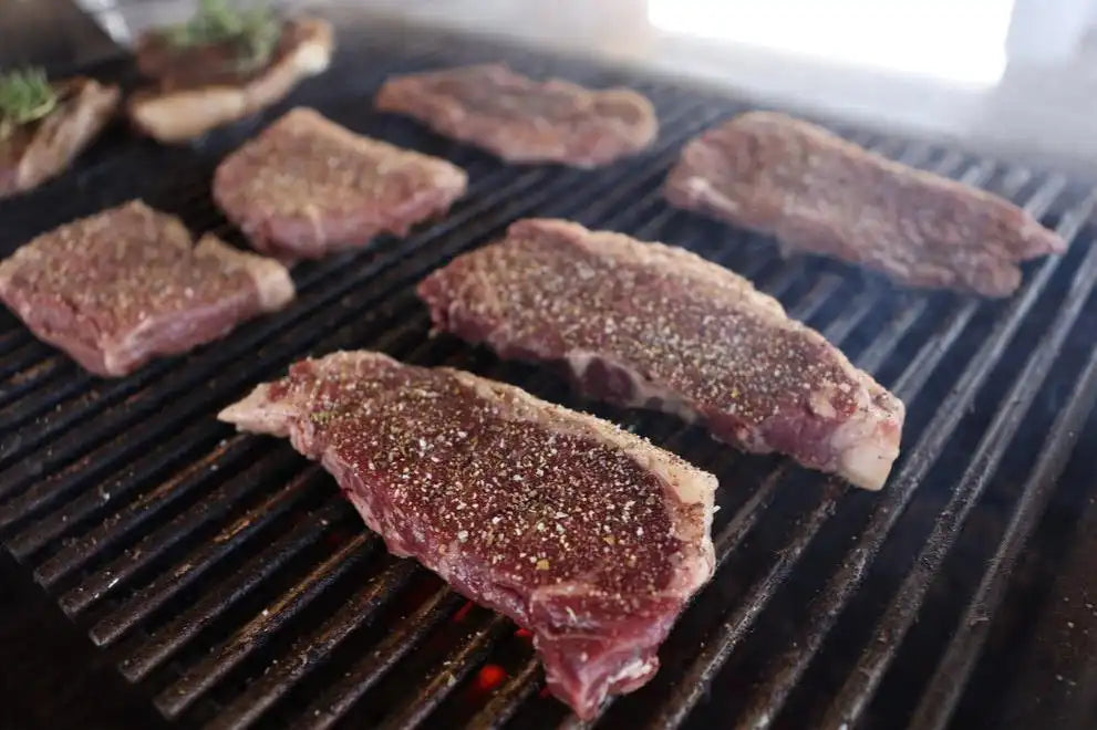 Thick-cut seasoned steaks cooking on a grill with visible smoke, capturing the rich texture and flavor of Drummond Ranch’s premium cuts.