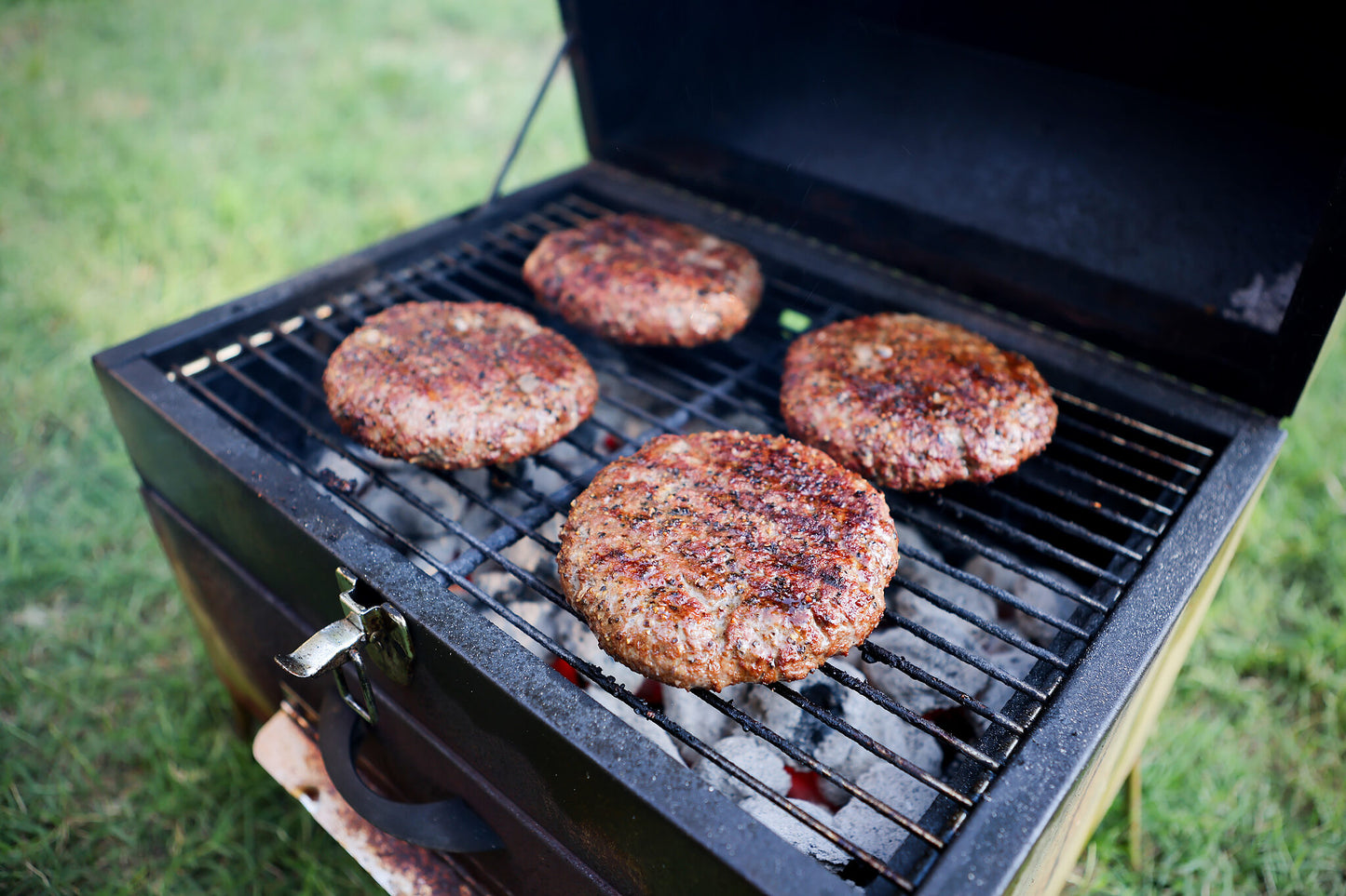 Four large, seasoned burger patties grilling over hot coals on a charcoal grill, with a grassy background setting the scene for a classic cookout.