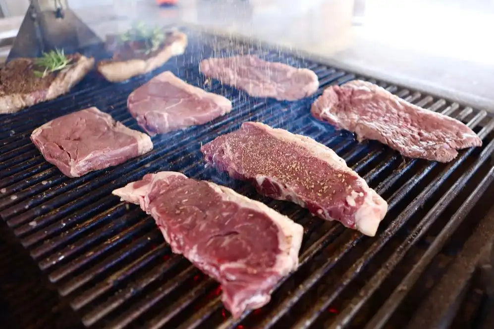 A grill full of assorted raw steaks being seasoned mid-cook, showcasing the marbling and preparation of Drummond Ranch beef for a flavorful result.