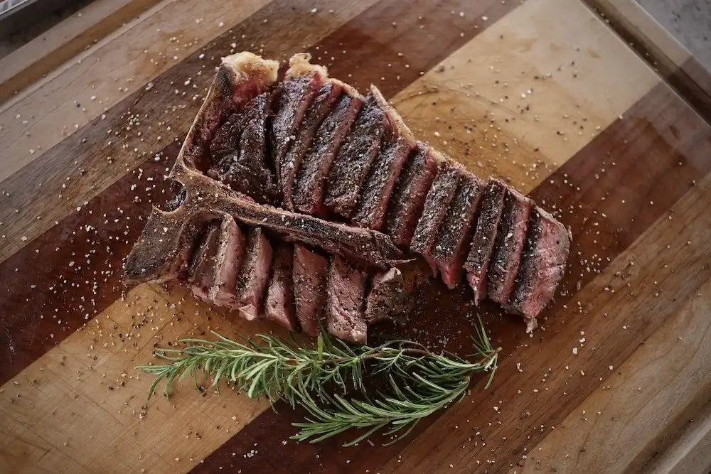 Perfectly grilled, sliced T-bone steak seasoned with salt and pepper, displayed on a wooden board with a sprig of rosemary for garnish.