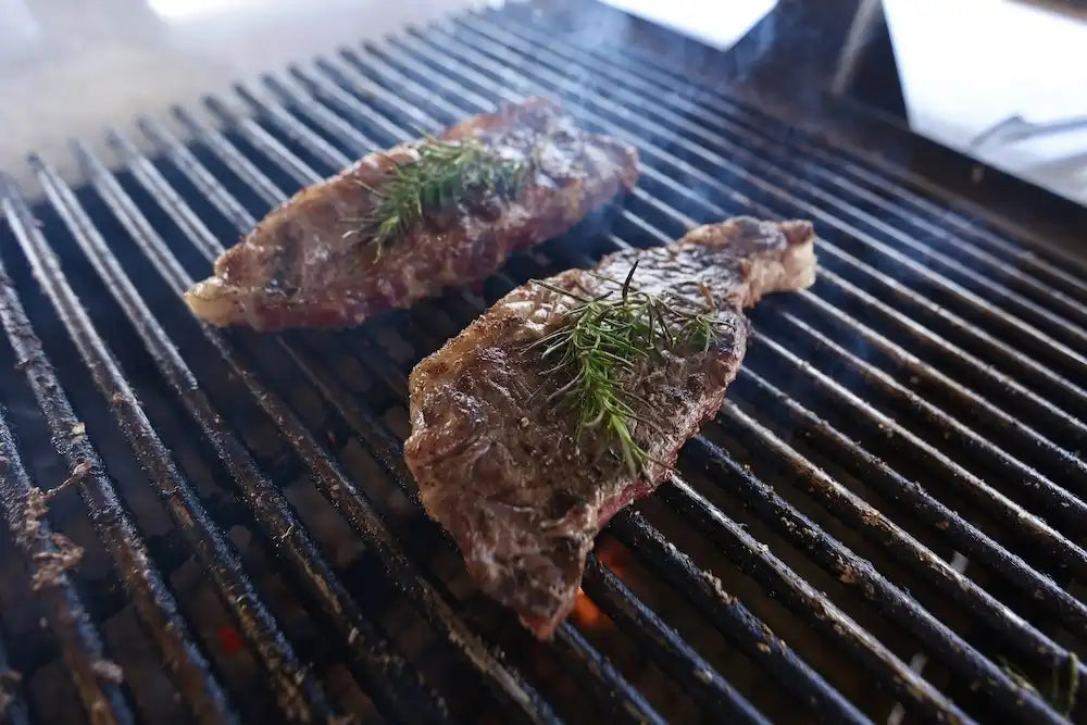 Two seasoned steaks topped with fresh rosemary grilling over open flames, capturing the flavor and simplicity of Drummond Ranch beef.