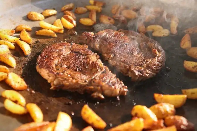 Two seared steaks sizzling on a flat-top grill surrounded by golden-brown potato wedges, showcasing a hearty Drummond Ranch meal in progress.