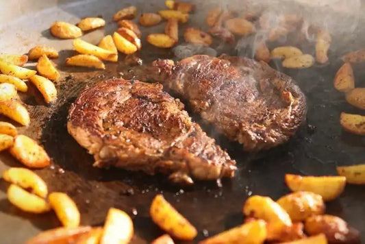 Two seared steaks sizzling on a flat-top grill surrounded by golden-brown potato wedges, showcasing a hearty Drummond Ranch meal in progress.