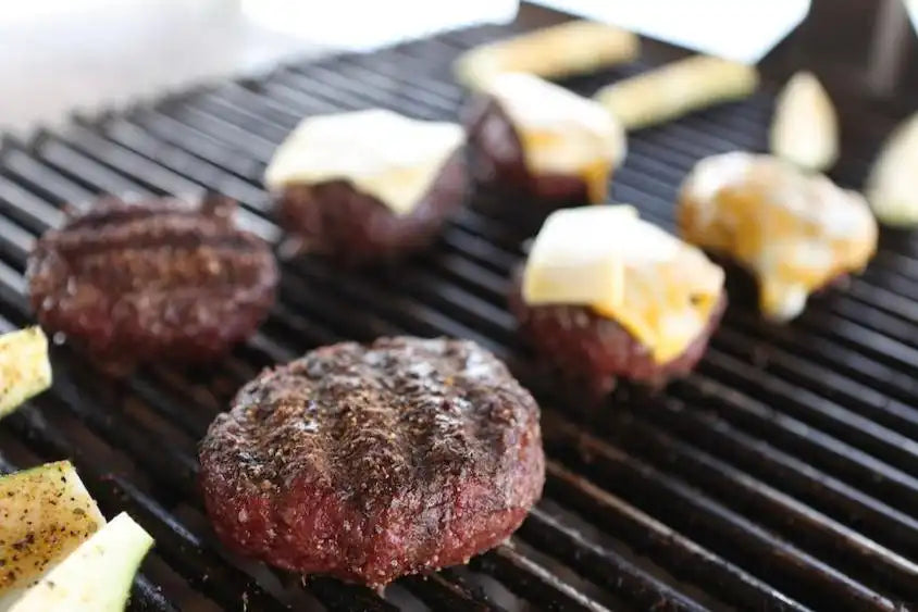A mix of cheeseburgers and plain beef patties cooking on a grill with sliced vegetables, representing the full-flavored results of Drummond Ranch ground beef.