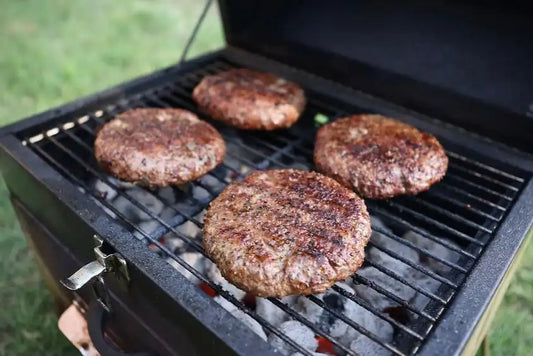 Four well-seasoned hamburger patties cooking over hot coals in a charcoal grill, ready to serve with that classic backyard barbecue flavor.