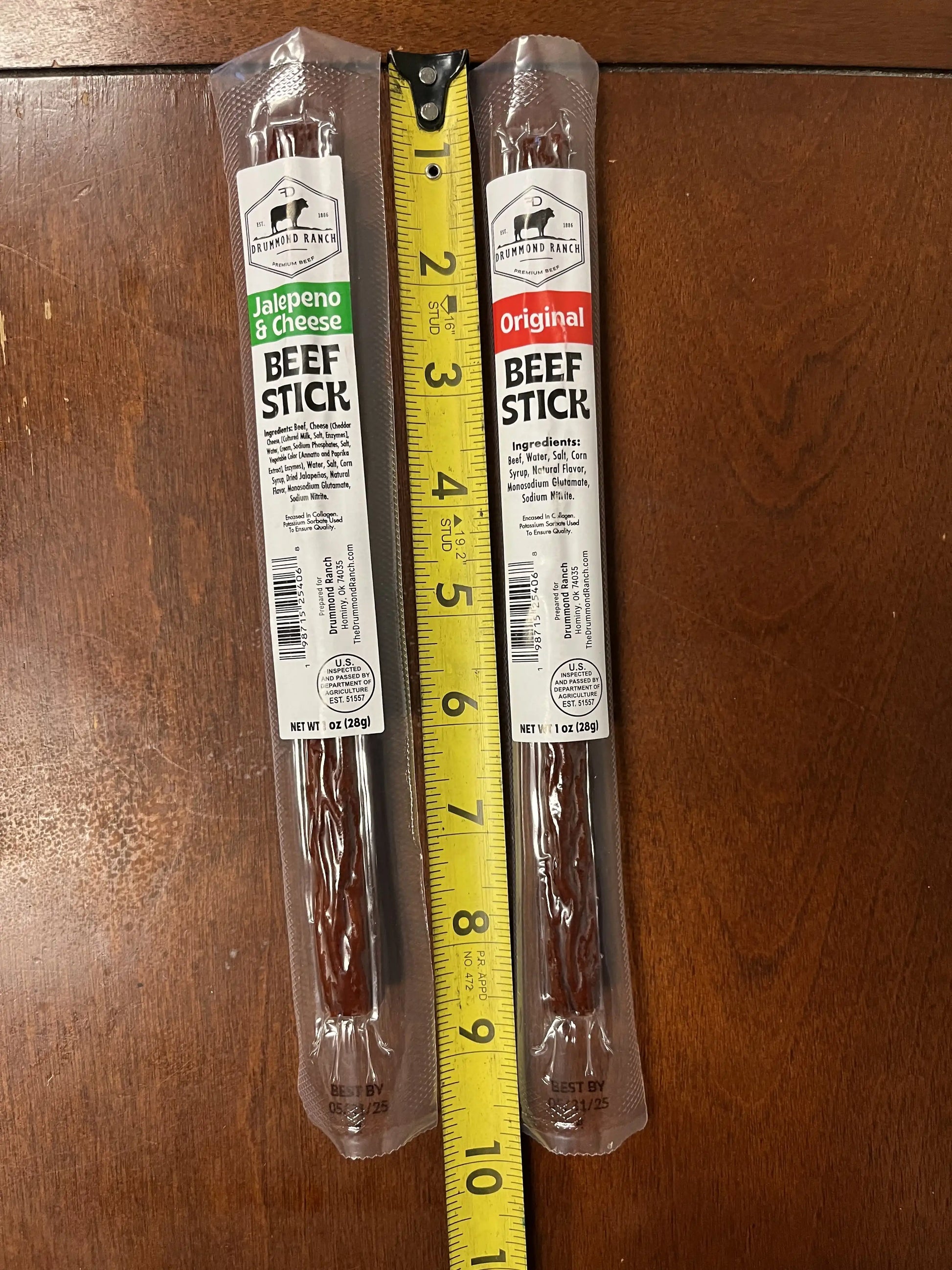 Two individually packaged Drummond Ranch beef sticks, one Original and one Jalapeño & Cheese, placed side-by-side next to a measuring tape showing their length.