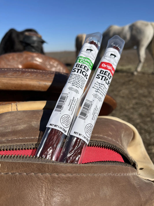 Original and Jalapeño & Cheese beef sticks from Drummond Ranch sticking out of a leather saddle bag, with horses grazing in the background.