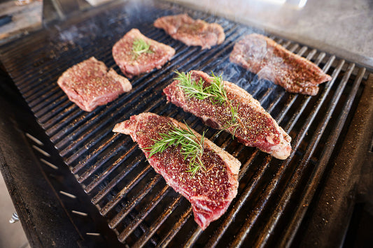 Close-up of seasoned raw steaks, including ribeyes, on a hot grill with visible smoke and rosemary, showcasing Drummond Ranch’s premium grilling cuts.