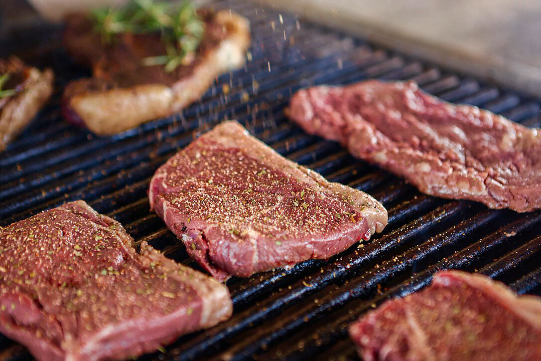 Raw, well-marbled steaks sizzling on the grill as seasoning is sprinkled on top—capturing the freshness and care in preparing Drummond Ranch beef.
