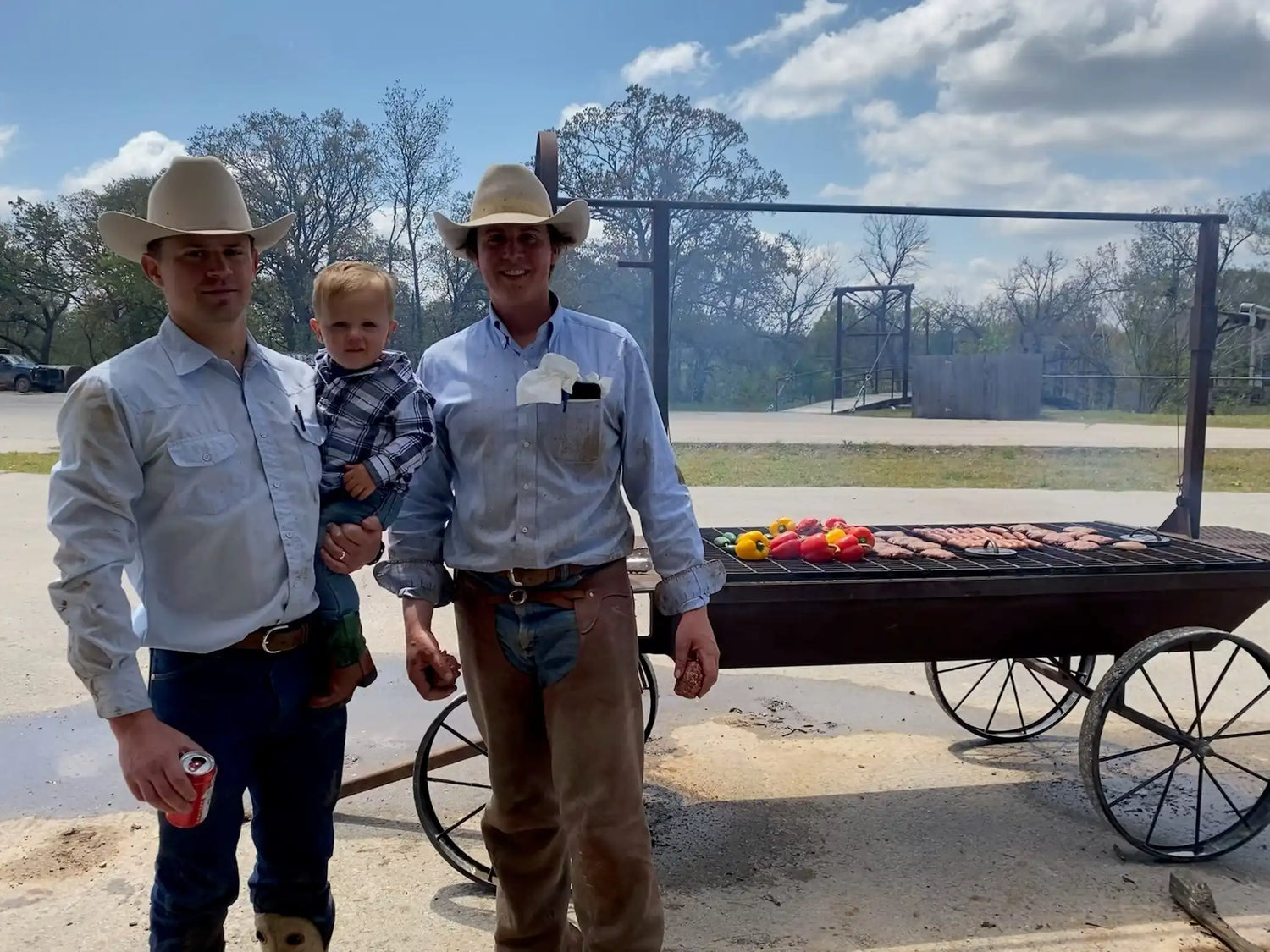 Two cowboys and a young child standing beside a custom grill filled with colorful peppers and beef cuts, sharing a moment during outdoor cooking at Drummond Ranch.