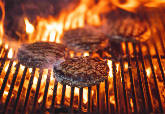Juicy hamburger patties grilling over an open flame, with smoke rising from the fire, showcasing Drummond Ranch’s rich, flavorful ground beef.