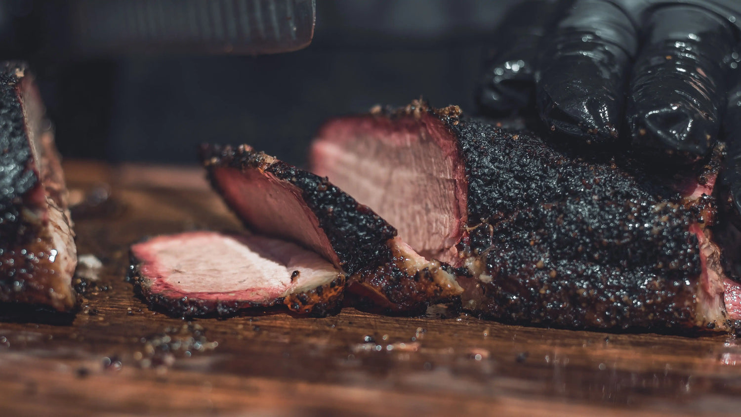 Sliced smoked beef brisket on a wooden cutting board, showing a thick pink smoke ring and bark, with gloved hands holding the meat.