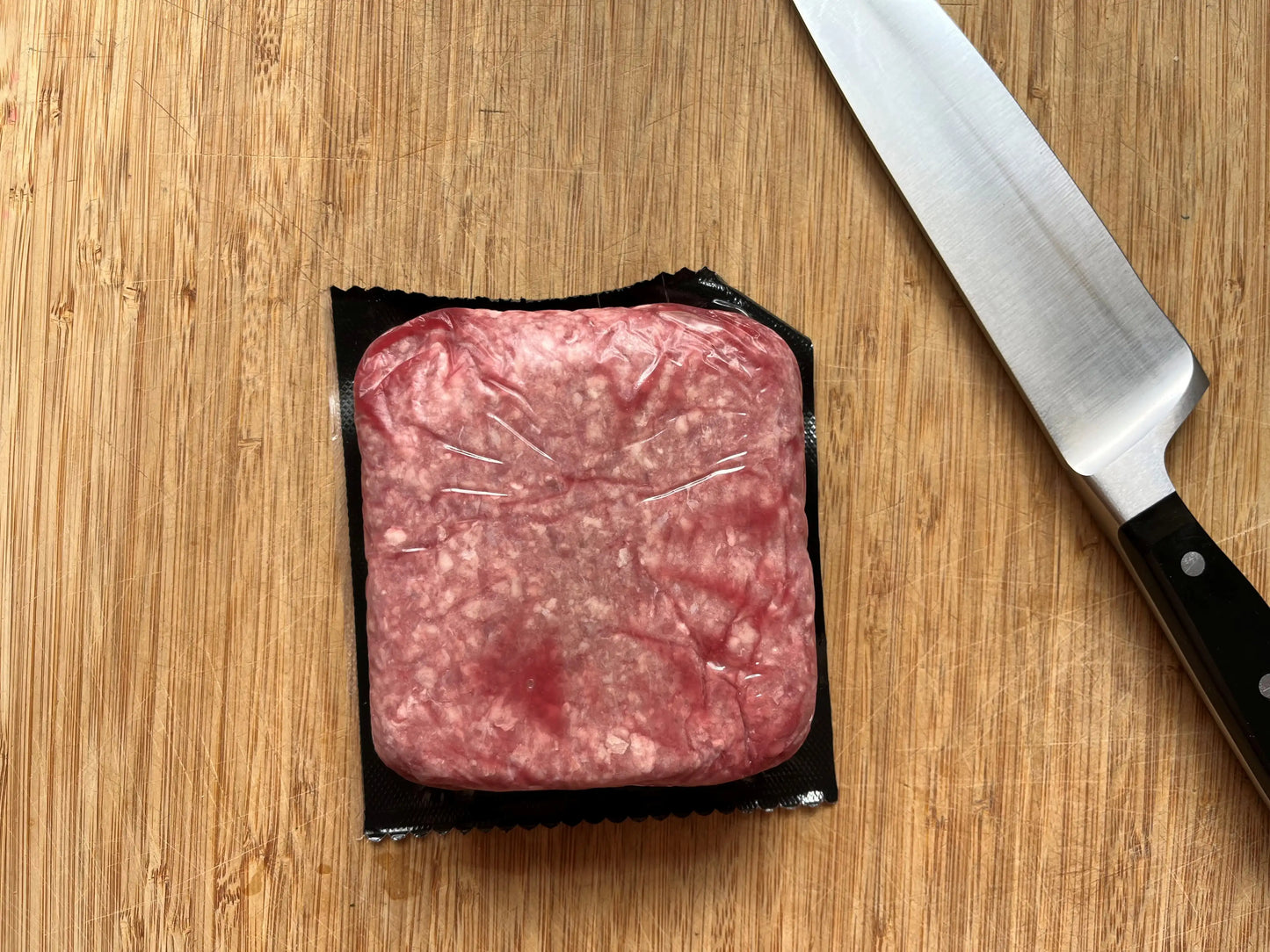 Vacuum-sealed square package of raw ground beef from Drummond Ranch on a wooden cutting board, next to a chef’s knife.