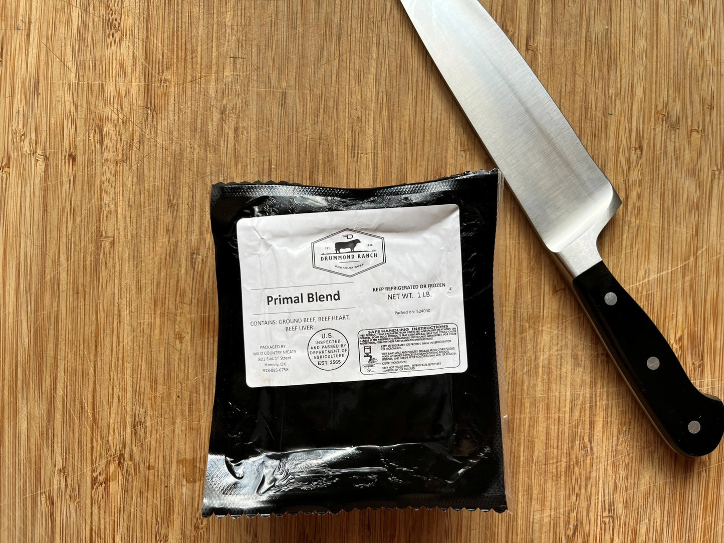 A sealed one-pound package of Drummond Ranch Primal Blend ground beef on a wooden cutting board next to a large kitchen knife.