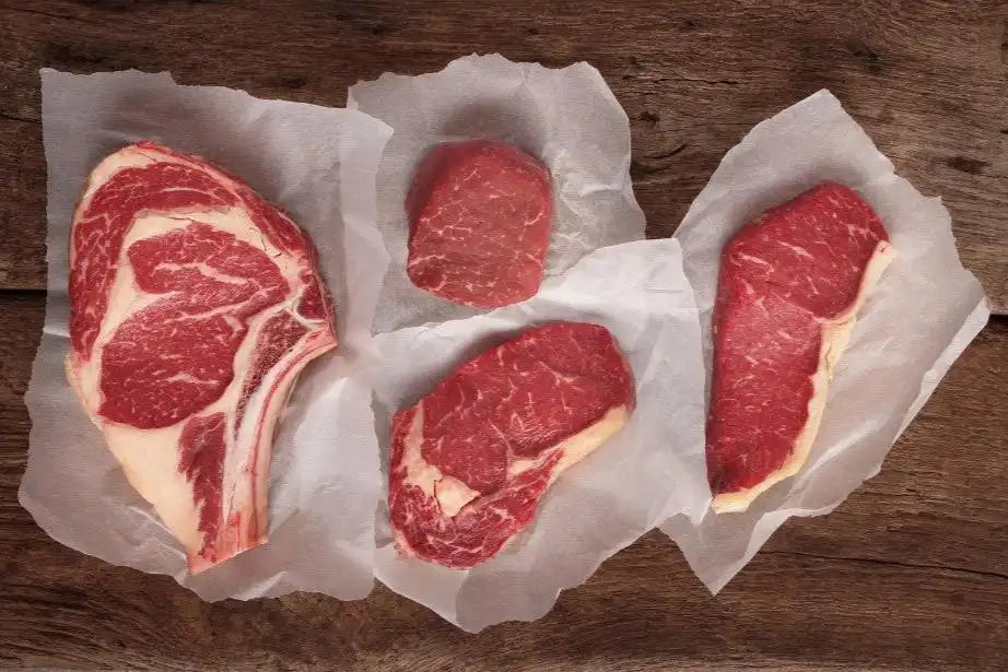 Four cuts of raw beef—bone-in ribeye, filet, strip, and sirloin—neatly displayed on parchment paper over a rustic wood surface, highlighting variety in Drummond Ranch steaks.