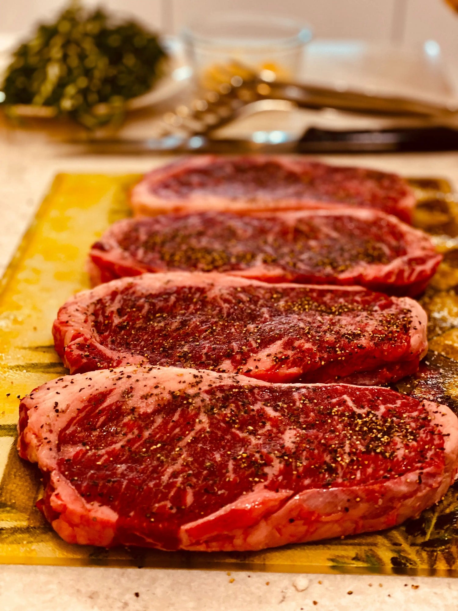 Four raw ribeye steaks from Drummond Ranch, seasoned with coarse black pepper, displayed on a yellow cutting board in a home kitchen setting.