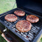 Four large, seasoned burger patties grilling over hot coals on a charcoal grill, with a grassy background setting the scene for a classic cookout.